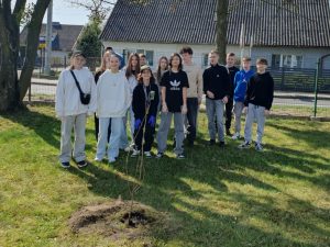 Grupa starszych uczniów stoi na trawie za posadzonym drzewkiem podczas szkolnej akcji sadzenia.