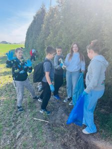 Uczniowie wraz z nauczycielem zbierają odpady przy zadrzewieniu w pobliżu szkoły, używając rękawic i worków na śmieci.