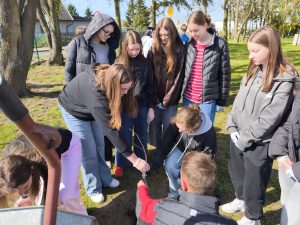 Grupa starszych uczennic sadzi drzewko; jedna osoba umieszcza sadzonkę w ziemi, pozostałe obserwują i pomagają.