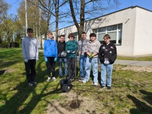 Grupa uczniów stoi na trawie przy posadzonym drzewku; przed nimi widoczna jest sadzonka i wiadro z wodą.