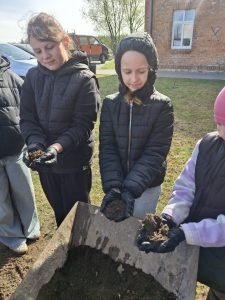 Uczennice trzymają w dłoniach ziemię przygotowaną do sadzenia roślin; przed nimi taczka z podłożem.