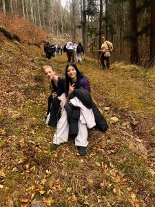 Dwie uczennice siedzą na leśnej ścieżce, a grupa uczniów z nauczycielem idzie w tle podczas wycieczki.