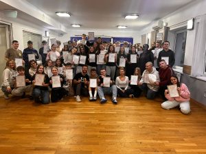 Uczniowie klasy 8 i opiekunowie stoją i klęczą w sali, trzymając certyfikaty ukończenia zajęć EuroWeek.