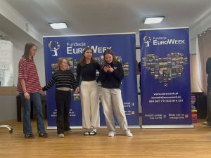 Trzy uczennice prezentują krótką scenkę na scenie przed banerami EuroWeek podczas zajęć językowych.