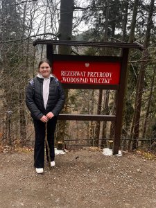Uczennica stoi przy tablicy informacyjnej rezerwatu „Wodospad Wilczki” podczas wycieczki terenowej.