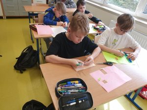 Uczniowie siedzą w klasie przy ławkach i pracują nad makietami. Wycinają z papieru figury geometryczne i przygotowują elementy zabudowy miasta.