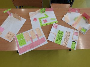 Na ławkach szkolnych znajdują się papierowe makiety miasteczka wykonane przez uczniów. Widać budynki, drogi oraz elementy przestrzenne wykonane z kolorowego papieru. Projekty przedstawiają układ miasta zbudowany z figur geometrycznych – głównie kwadratów i prostokątów.