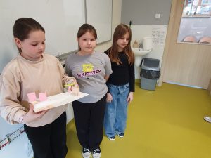 Trzy uczennice stoją w klasie. Jedna z nich trzyma makietę przedstawiającą fragment miasteczka z papierowymi budynkami i drogą. Uczennice prezentują swoją pracę wykonaną podczas zajęć matematycznych metodą STEAM.