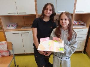 Dwie uczennice stoją w klasie i prezentują makietę miasta wykonaną z papieru. Na makiecie znajdują się budynki, droga oraz elementy przestrzenne wykonane z kolorowego papieru.