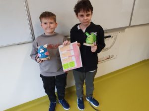 Dwóch uczniów stoi w klasie i prezentuje swoje prace. Jeden trzyma makietę miasta z papieru, drugi przestrzenną figurę wykonaną z kolorowych materiałów.
