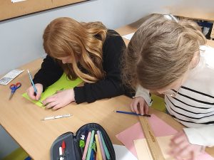 Uczennice siedzą przy ławce i rysują oraz wycinają elementy z kolorowego papieru przy użyciu linijki i nożyczek. Przygotowują elementy do makiety miasta.