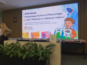 Sala z ekranem prezentującym informacje o konkursie oraz stołem z przygotowanymi nagrodami.