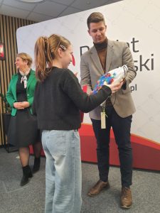 Uczennica odbiera nagrodę rzeczową od organizatora podczas ceremonii konkursowej.
