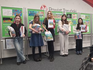Uczennice stoją przed wystawą prac plastycznych i trzymają dyplomy oraz nagrody w konkursie plastycznym.