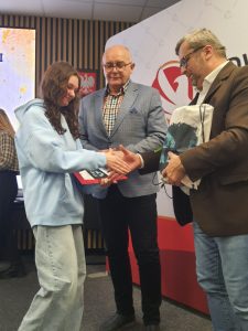 Uczennica odbiera dyplom i nagrodę od organizatorów podczas uroczystości konkursowej.