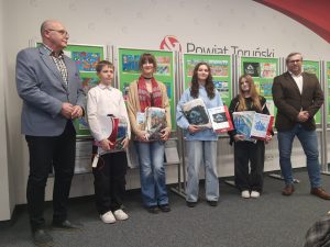 Laureaci konkursu plastycznego wraz z organizatorami pozują do zdjęcia na tle wystawy prac.