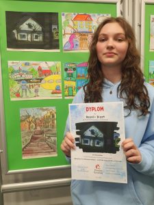 Uczennica trzyma dyplom za zwycięstwo w konkursie plastycznym, stojąc przy wystawie prac.