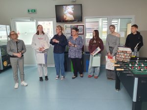 Uczniowie i nauczyciele stoją w szkolnym holu podczas wręczenia nagród w konkursie literackim; laureaci trzymają książki i torby z upominkami.