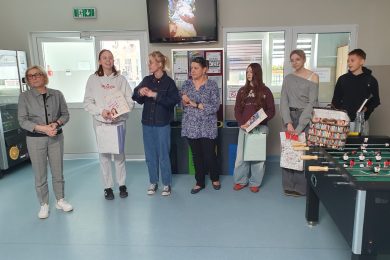 Uczniowie i nauczyciele stoją w szkolnym holu podczas wręczenia nagród w konkursie literackim; laureaci trzymają książki i torby z upominkami.