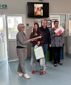 Uczennica odbiera nagrodę książkową, stojąc między nauczycielkami w szkolnym holu