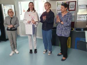 „Uczennica odbiera nagrodę książkową za najlepszą fraszkę; obok nauczycielki biją brawo.”