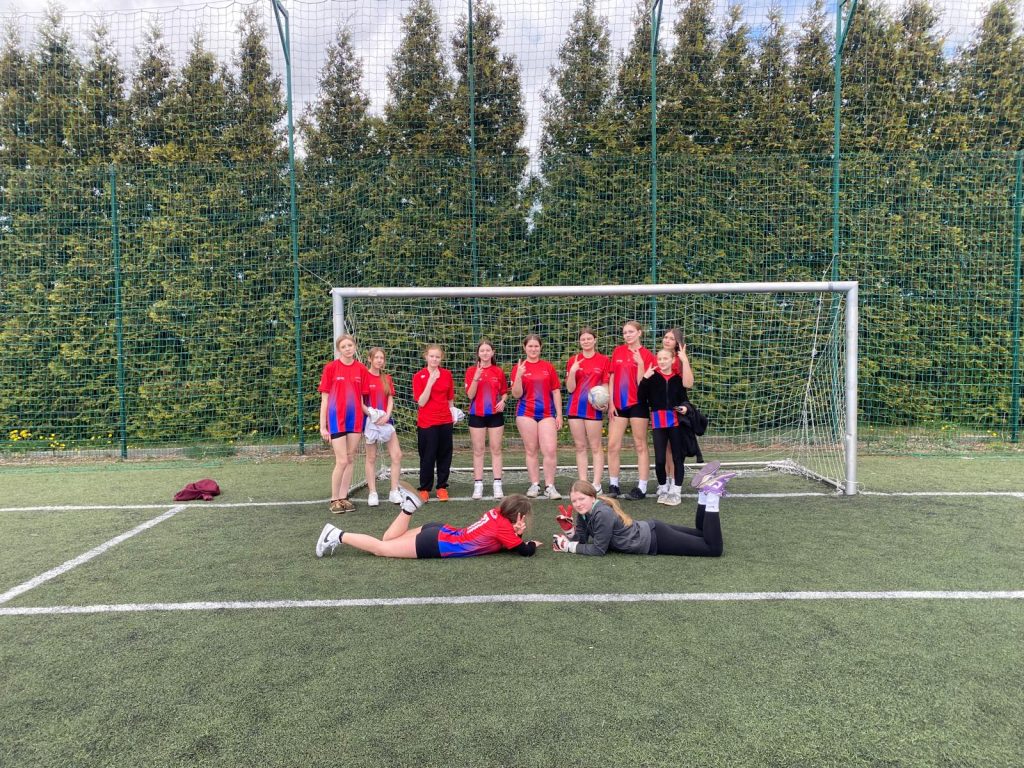 Grupa młodych dziewcząt w czerwono-niebieskich koszulkach sportowych stoi na boisku piłkarskim przed bramką z siatką. Dwie dziewczyny leżą na murawie na pierwszym planie, uśmiechając się i pozując. W tle widoczne są wysokie drzewa oraz ogrodzenie z siatki.