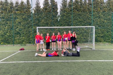 Grupa młodych dziewcząt w czerwono-niebieskich koszulkach sportowych stoi na boisku piłkarskim przed bramką z siatką. Dwie dziewczyny leżą na murawie na pierwszym planie, uśmiechając się i pozując. W tle widoczne są wysokie drzewa oraz ogrodzenie z siatki.