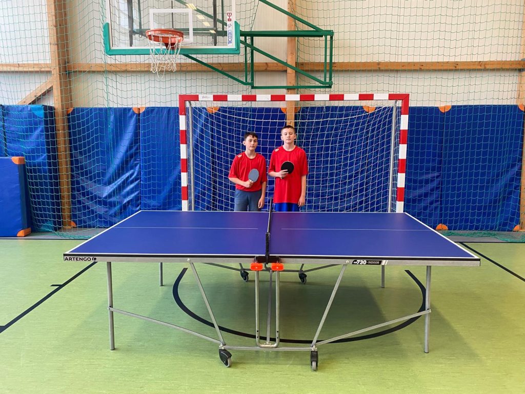 Dwóch chłopców stoi za stołem do tenisa stołowego w sali gimnastycznej. Obaj trzymają rakietki i mają na sobie czerwone koszulki. W tle widoczna jest bramka do piłki ręcznej oraz kosz do koszykówki zawieszony nad nią.