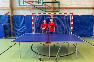 Dwóch chłopców stoi za stołem do tenisa stołowego w sali gimnastycznej. Obaj trzymają rakietki i mają na sobie czerwone koszulki. W tle widoczna jest bramka do piłki ręcznej oraz kosz do koszykówki zawieszony nad nią.