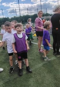 Dzieci z oddziału przedszkolnego oczekują na rozpoczęcie konkurencji sportowych podczas Olimpiady Przedszkolaka. Uczestnicy ubrani w stroje sportowe stoją wraz z opiekunami na boisku wielofunkcyjnym.