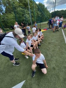 Dzieci siedzą na ławce i przygotowują się do kolejnych konkurencji sportowych. Obok znajduje się nauczycielka wspierająca uczestników zawodów. W tle widoczne są inne drużyny oraz opiekunowie.