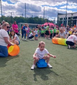 Przedszkolak bierze udział w konkurencji sprawnościowej, przemieszczając się na plastikowym sprzęcie sportowym. W tle odbywają się kolejne konkurencje z wykorzystaniem kolorowych tuneli i przyborów sportowych.