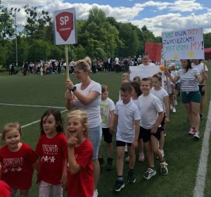 Uczestnicy oddziału przedszkolnego Szkoły Podstawowej w Ostaszewie maszerują podczas uroczystego otwarcia VIII Olimpiady Przedszkolaka „Jelonki na start”. Dzieci niosą tabliczkę z nazwą szkoły oraz transparent dopingujący drużynę. W tle widoczni są pozostali uczestnicy wydarzenia zgromadzeni na boisku.
