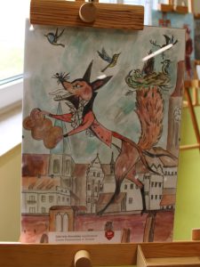 Praca przedstawia lisa w fantazyjnym stroju spacerującego po mieście. W tle znajdują się kamienice i drzewo z ptasim gniazdem. Ilustracja wykonana została w stylu bajkowym, z wykorzystaniem ciepłych odcieni brązu i pomarańczu.