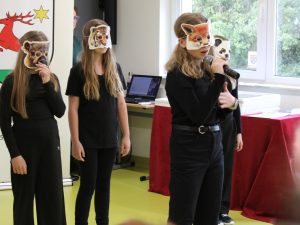 Uczennice podczas występu recytatorskiego. Jedna z uczestniczek recytuje tekst do mikrofonu, pozostałe występują w maskach zwierząt.