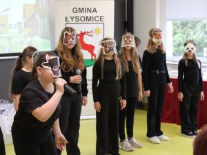 Fragment występu recytatorskiego uczestników konkursu. Dzieci w czarnych strojach prezentują program artystyczny w maskach przedstawiających zwierzęta.