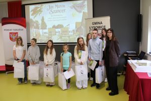 Grupa laureatów konkursu wraz z organizatorami podczas wręczenia nagród. Uczniowie stoją na scenie z dyplomami i upominkami. W tle ekran z grafiką poświęconą Janowi Marcinowi Szancerowi oraz banery partnerów wydarzenia.
