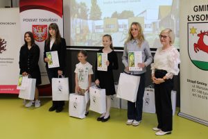 Uczestnicy konkursu prezentują dyplomy i nagrody podczas uroczystego rozdania wyróżnień. W tle widoczna prezentacja poświęcona Janowi Marcinowi Szancerowi.