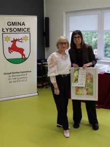 Wręczenie podziękowania dla osoby zaangażowanej w organizację konkursu. Dwie osoby prezentują oprawioną pracę plastyczną i dyplom.