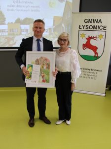 Przedstawiciel samorządu i organizatorka konkursu prezentują podziękowanie oraz oprawioną pracę plastyczną.