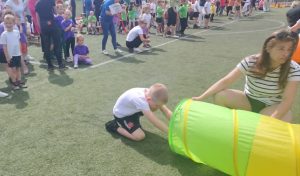 Dziecko uczestniczy w konkurencji sportowej podczas Olimpiady Przedszkolaka na boisku szkolnym. Chłopiec przechodzi przez kolorowy tunel, a obok nauczycielka wspiera przebieg zadania. W tle widoczne są drużyny przedszkolne oczekujące na swoją kolej oraz opiekunowie obserwujący zawody.