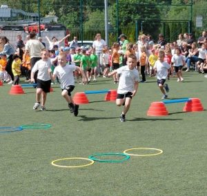 Dzieci biorą udział w biegu sztafetowym podczas konkurencji sportowej. Uczestnicy pokonują wyznaczoną trasę z wykorzystaniem obręczy i przeszkód ustawionych na boisku. W tle widoczne są drużyny kibicujące zawodnikom.