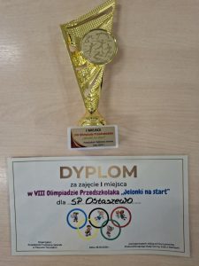 Puchar i dyplom za zajęcie I miejsca zdobyte przez Szkołę Podstawową w Ostaszewie podczas VIII Olimpiady Przedszkolaka „Jelonki na start”. Na dyplomie widnieje informacja o zwycięstwie oraz nazwa placówki.