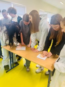 Zdjęcie przedstawia uczniów klas VII–VIII uczestniczących w praktycznych ćwiczeniach dotyczących samobadania piersi. Młodzież wykonuje ćwiczenia przy użyciu fantomów umieszczonych na stole.