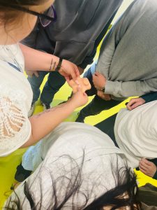 Zdjęcie przedstawia prowadzącą zajęcia demonstrującą uczniom sposób wykonywania samobadania na modelu edukacyjnym. Uczniowie stoją wokół stołu i obserwują pokaz praktyczny.