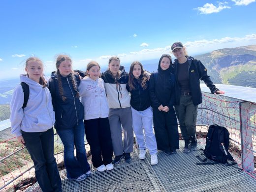 Grupa uczennic wraz z opiekunką stoi na platformie widokowej na Śnieżce. W tle rozciąga się panorama gór i dolin.