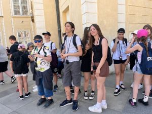 Grupa uczniów podczas zwiedzania Pragi stoi przed zabytkowym budynkiem. Uczestnicy słuchają informacji przekazywanych przez przewodnika za pomocą systemu słuchawkowego.