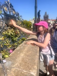 Uczennica dotyka płaskorzeźby przedstawiającej psa na balustradzie Mostu Karola w Pradze. W tle widoczne zabytkowe budowle miasta.