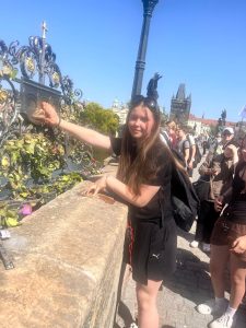 Uczennica pozuje przy płaskorzeźbie psa na Moście Karola. W tle znajdują się spacerujący turyści oraz panorama historycznego centrum Pragi.