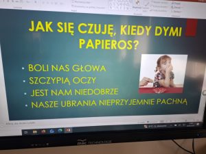 Slajd prezentacji edukacyjnej wyświetlony podczas zajęć programu. Materiał przedstawia skutki przebywania w zadymionym otoczeniu, takie jak ból głowy, szczypanie oczu i nieprzyjemny zapach ubrań.
